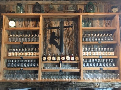 A wooden shelf displays numerous bottles and glasses against a rustic backdrop, featuring a silhouette of a person climbing.