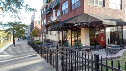 Outdoor seating area of Fresco Italian Café beside a canal, with a brick building and trees lining the walkway.