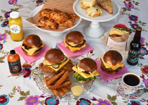 A colorful spread of burgers, pastries, sauces, and drinks on a floral tablecloth.