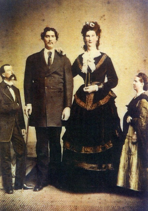 A vintage photo of four people, including a tall man and a woman in a black dress, posing together in 19th-century attire.