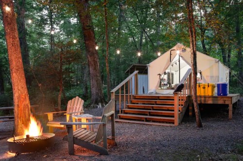 A cozy campsite with a tent, wooden deck, fire pit, and string lights in a lush forest setting.