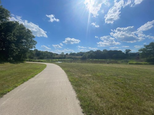 A sunny park scene with a winding path, green grass, trees, and a pond under a blue sky with fluffy clouds.