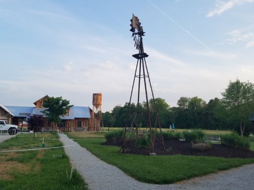 A rustic landscape featuring a windmill, gravel path, and a wooden barn under a clear sky. Green trees in the background.