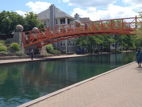 An orange bridge spans a calm canal, with greenery and buildings in the background on a sunny day.