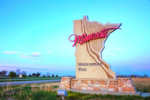 “Minnesota Welcomes You” roadside sign featuring the state outline and text, positioned near a highway surrounded by trees and grass.