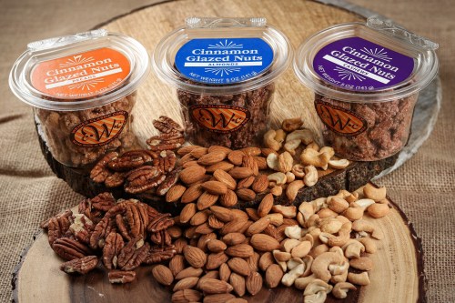 Three containers of cinnamon glazed nuts (pecans, almonds, and mixed nuts) arranged on a wooden surface with scattered nuts.