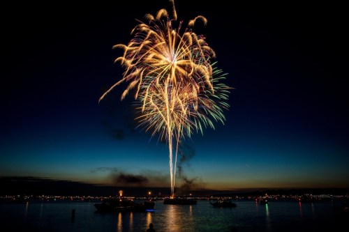 Colorful fireworks burst over a calm body of water at night, illuminating the sky and reflecting on the surface.