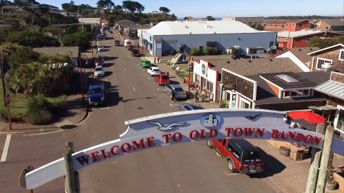 A view of Old Town Bandon, featuring shops, cars, and a welcoming sign against a clear sky.