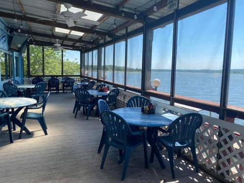 A spacious outdoor dining area with blue tables and chairs overlooking a calm lake, surrounded by screens.