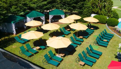Aerial view of a grassy area with green lounge chairs, yellow umbrellas, and tents, surrounded by trees and landscaping.