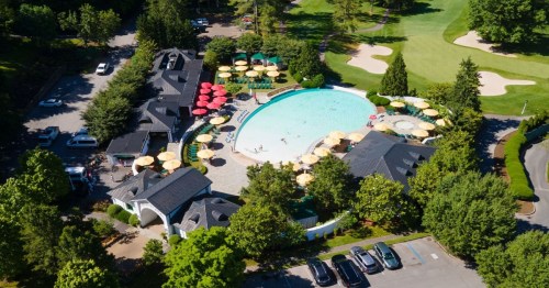 Aerial view of a resort pool surrounded by greenery, umbrellas, and a golf course in the background.