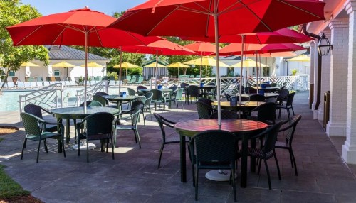 Outdoor poolside area with tables and red umbrellas, surrounded by lounge chairs and yellow umbrellas.