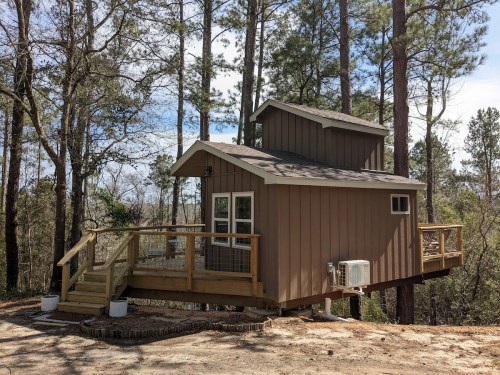 A small, modern cabin with a deck, surrounded by tall trees and a clear sky.