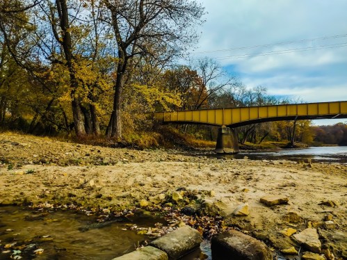A serene river scene with a yellow bridge, autumn trees, and a sandy bank under a cloudy sky.