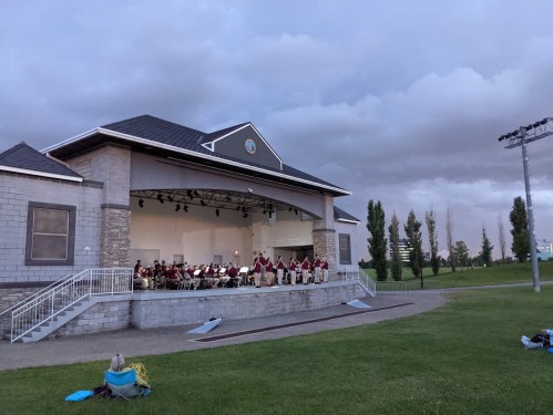 A band performs on an outdoor stage at dusk, with dark clouds overhead and green grass in the foreground.