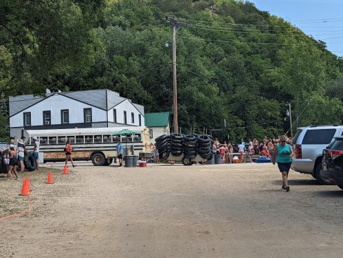 A busy outdoor scene with people near a bus and tire rentals, surrounded by greenery and a building in the background.