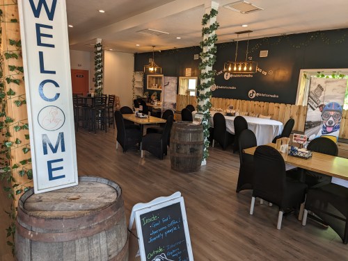 Cozy restaurant interior with tables, chairs, and a welcoming sign, featuring greenery and rustic decor.