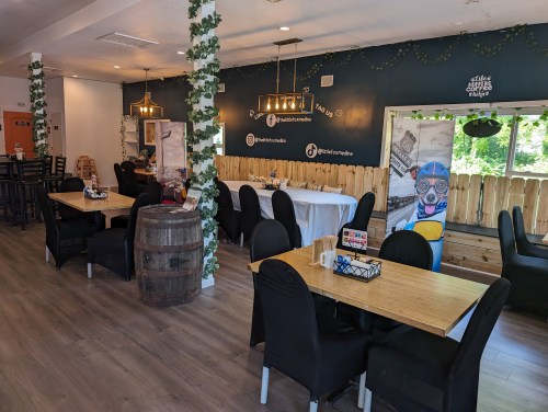 Cozy café interior with wooden tables, black chairs, and greenery decor, featuring a barrel and bright windows.