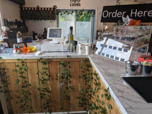 A cozy café counter with a marble top, cash register, and decorative plants, featuring a menu and display of pastries.