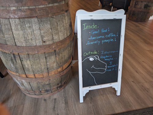 A chalkboard sign next to a wooden barrel, promoting great food and coffee, with a humorous note about raptors outside.