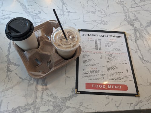 A coffee cup and iced drink on a marble table next to a menu for Little Fox Cafe & Bakery.