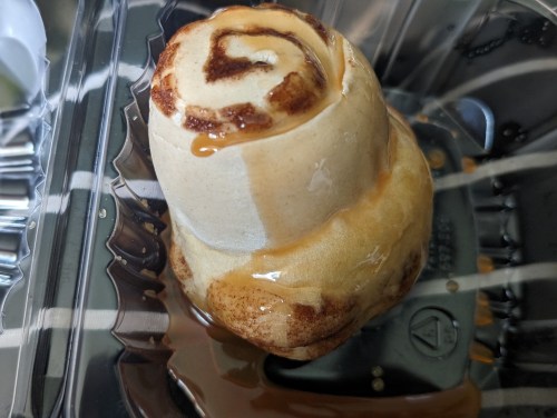 A cinnamon roll with a swirl design, topped with a glossy caramel glaze, placed in a clear plastic container.