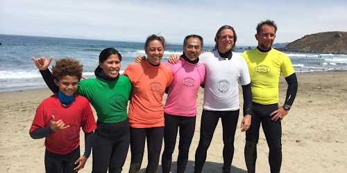 A group of six surfers in colorful wetsuits stands on the beach, smiling with the ocean in the background.