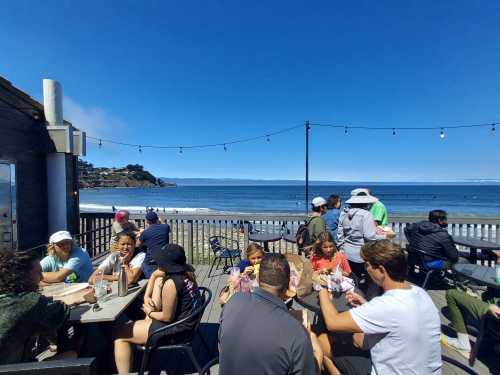 A lively outdoor dining area by the beach, with people enjoying food and a view of the ocean and surfers.