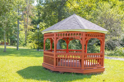 A red wooden gazebo with a peaked roof, surrounded by green grass and trees in a sunny park setting.