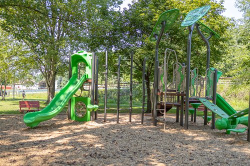 A green playground with a slide and climbing structures, surrounded by trees and a sandy area.