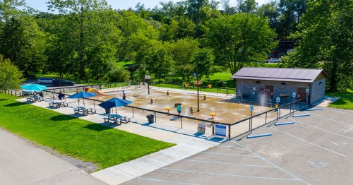 Aerial view of a splash pad with water features, picnic tables, and a restroom building surrounded by green trees.
