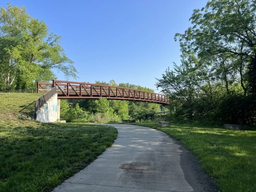 A wooden bridge arches over a path surrounded by lush greenery under a clear blue sky.