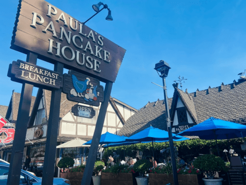 Sign for Paula's Pancake House with outdoor seating and blue umbrellas under a clear blue sky.