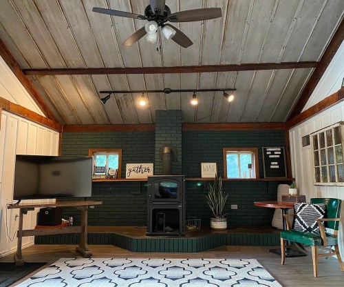 Cozy living room with a green brick fireplace, wooden ceiling, TV, and a small dining table with a green chair.