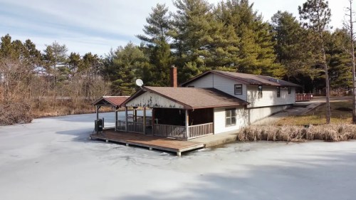A house on stilts by a frozen lake, surrounded by trees and a grassy area, with a deck extending over the ice.