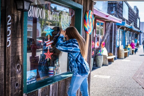 A girl peers into a candy shop window, admiring colorful treats and decorations on a sunny day.