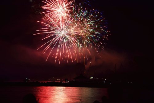 Colorful fireworks burst in the night sky over a calm lake, reflecting vibrant lights on the water's surface.