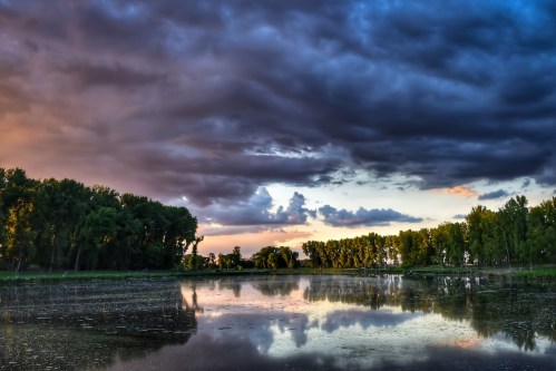 A serene river scene at sunset, with dark clouds and lush trees reflecting in the water.