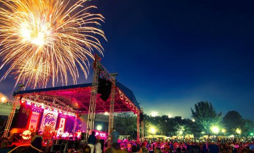 A lively outdoor concert scene with a large crowd enjoying fireworks in the night sky above the stage.
