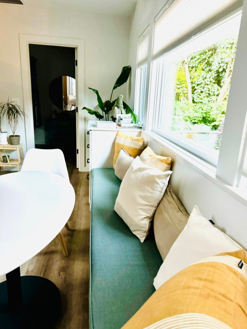 A cozy interior with a green bench, decorative pillows, a round table, and large windows overlooking greenery.