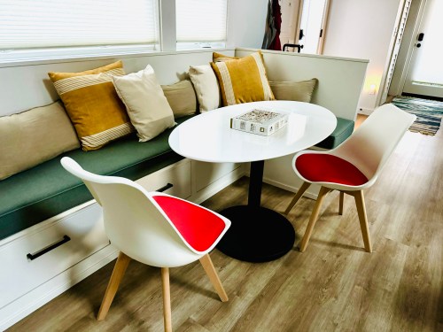 A cozy dining area featuring a round white table, two modern chairs, and a cushioned bench with colorful pillows.