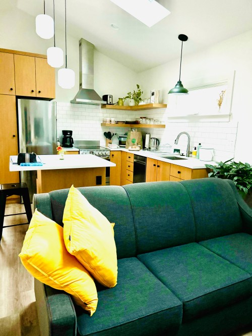 A modern kitchen with wooden cabinets, stainless steel appliances, and a cozy gray sofa with yellow pillows in the foreground.