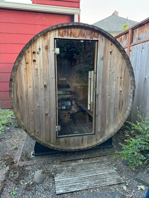 A round wooden sauna with a glass door, set in a garden area with gravel and plants.