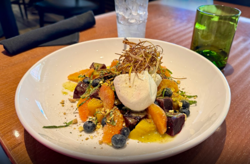 A colorful salad with beets, citrus, blueberries, and a scoop of ice cream, garnished with crispy greens on a white plate.