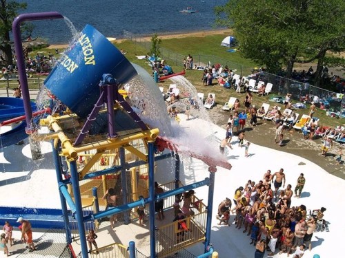 A water park scene with a large bucket pouring water over a crowd of people enjoying the attractions.