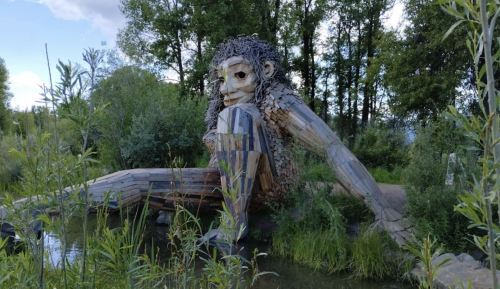 A large wooden sculpture of a seated figure surrounded by greenery and trees near a small body of water.