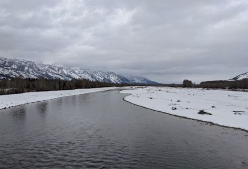 A winding river flows through a snowy landscape, with mountains in the background under a cloudy sky.