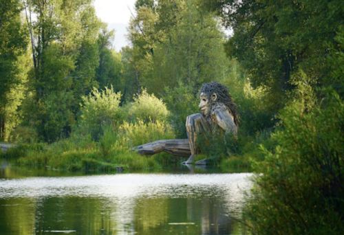 A large wooden sculpture of a seated figure with a lion-like mane near a tranquil lake surrounded by greenery.