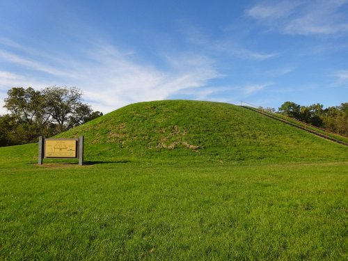 A grassy mound with a wooden sign in front, set against a blue sky and trees in the background.