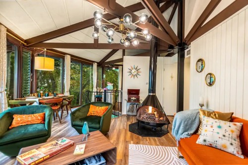 Cozy living room with modern decor, featuring a fireplace, colorful seating, and large windows overlooking nature.
